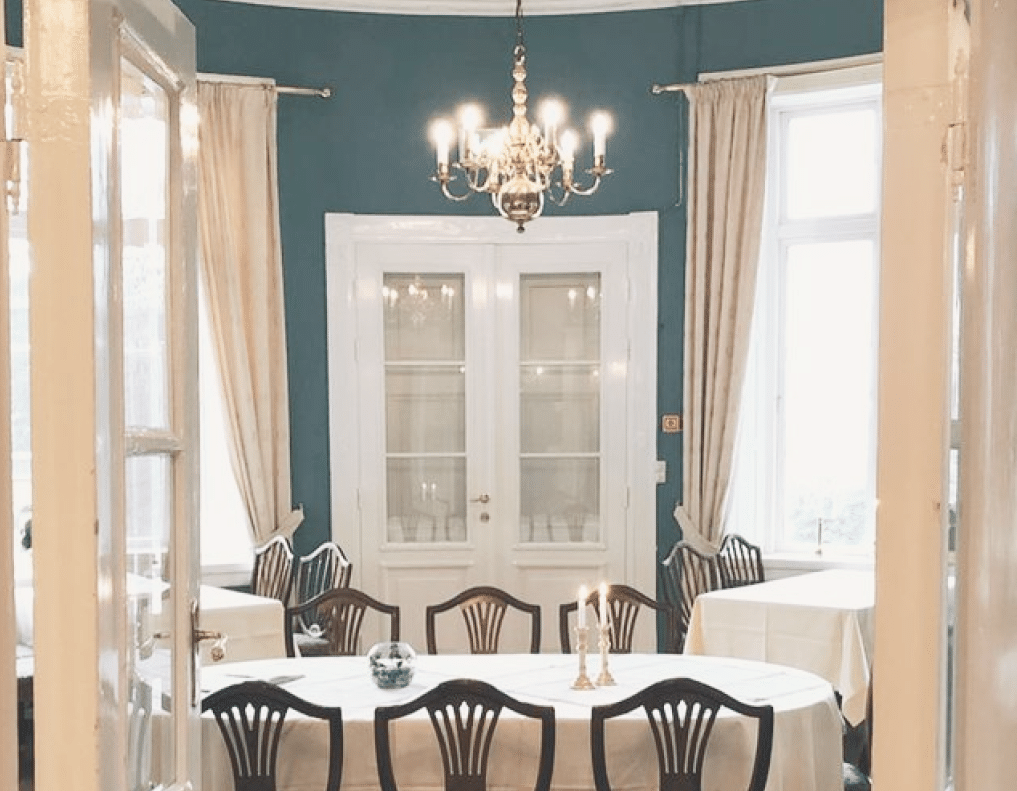 Elegant dining room with white curtains, a blue wall, a chandelier above a round table, surrounded by chairs, and double doors.